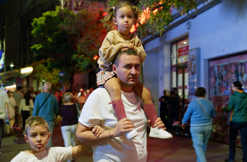 11 - A man in a white t-shirt carrying a young girl on his shoulders, with a boy standing next to them in a crowded evening street